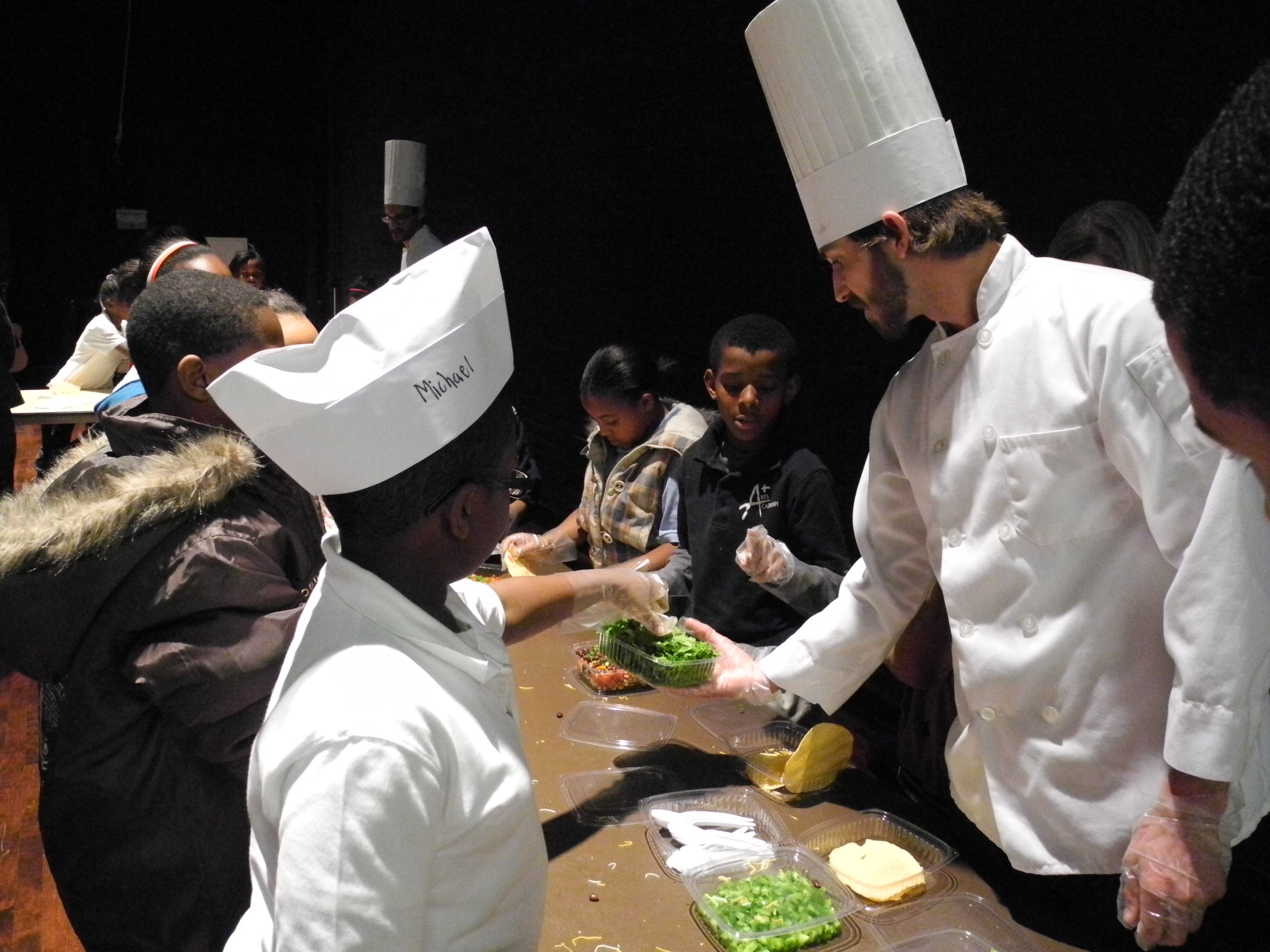 A chef holds a container of lettuce while students add ingredients to their meals.